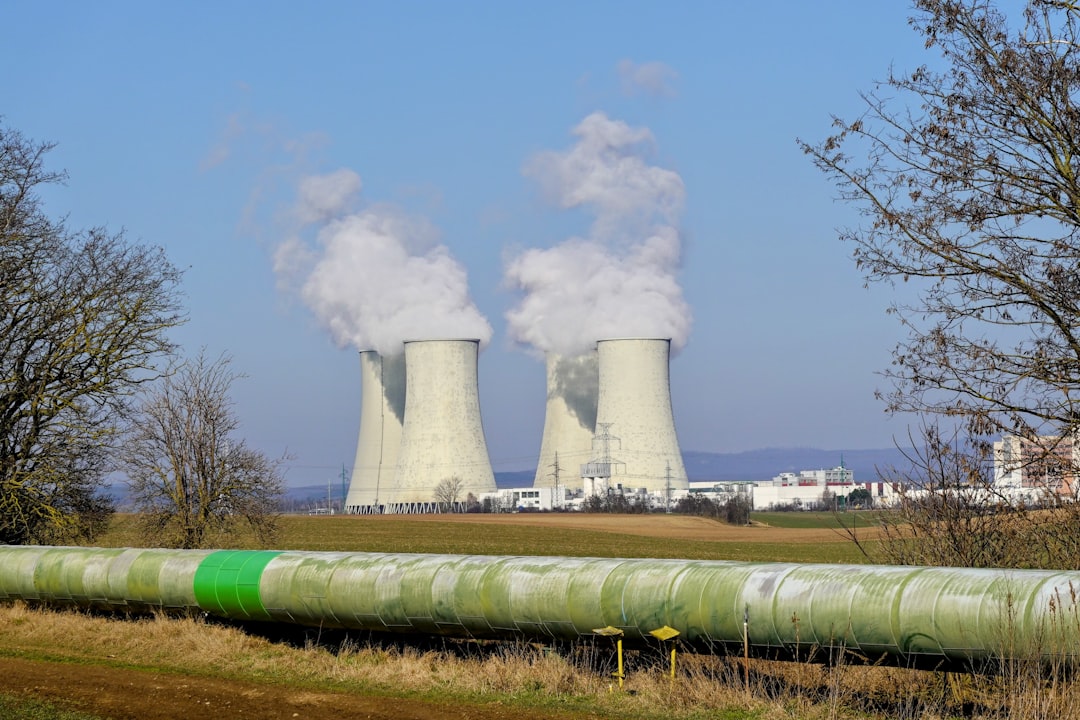 Wide landscape view of the Jaslovské Bohunice Nuclear Power Plant in Slovakia, with cooling towers releasing water vapor above a green field under a clear blue sky.