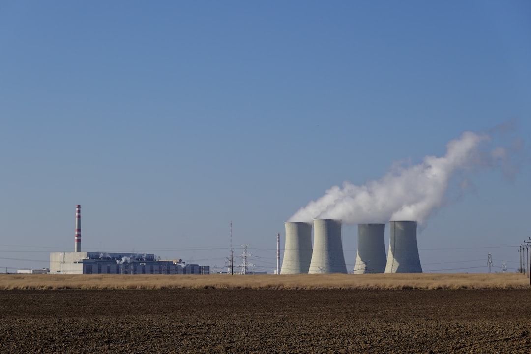 Wide landscape view of the Jaslovské Bohunice Nuclear Power Plant in Slovakia, with cooling towers releasing water vapor above a green field under a clear blue sky.